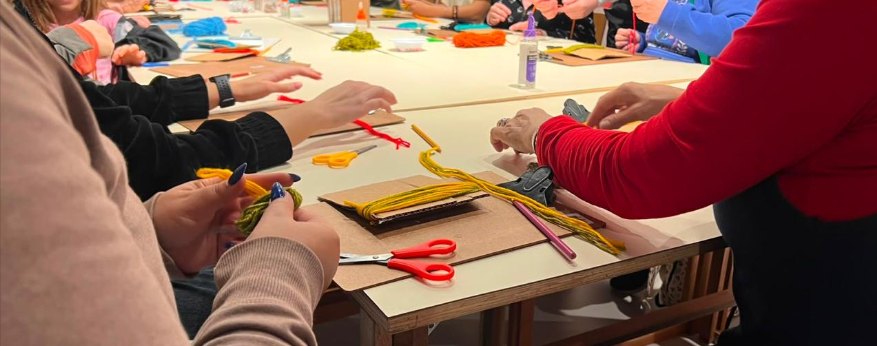 Em uma oficina da equipe educativa, muitas mãos ao redor de uma mesa comprida manuseiam fios de lã colorida, tesouras e papéis para criar bonecos encantados.